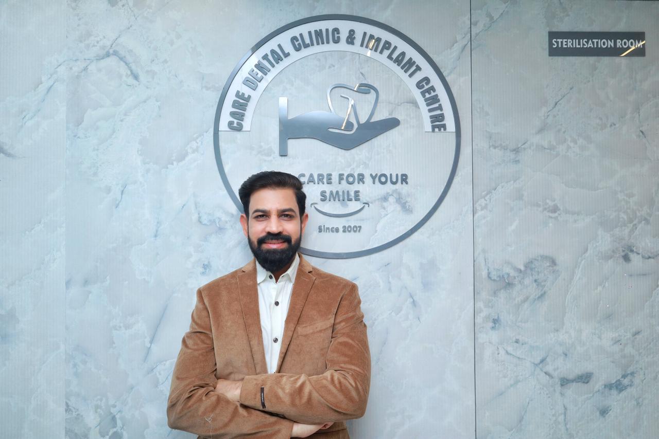 Dr. Gagan Jaiswal stands in front of a wall featuring logo of Care Dental Clinic and Implant Center in Indore.