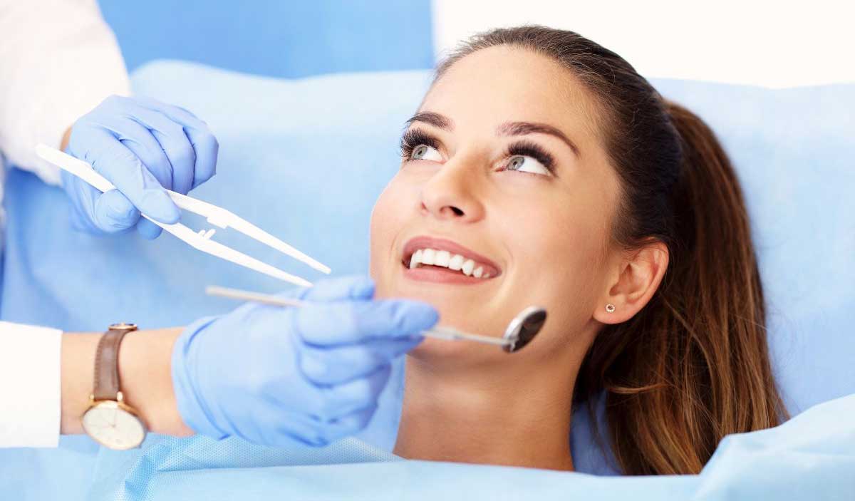 A woman smiling while a dentist cleans her teeth with a dental tool.
          